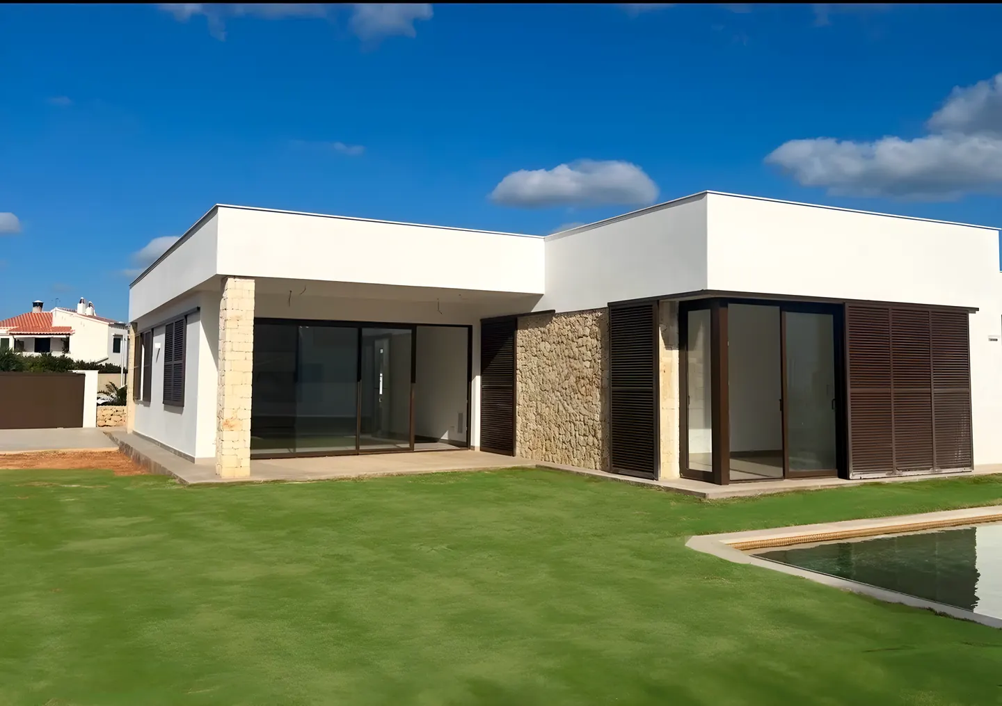 Casa de una planta en Menorca vista desde el jardín: volúmenes blancos con muro de piedra, grandes cristaleras con contraventanas de madera y piscina, bajo un cielo azul con nubes