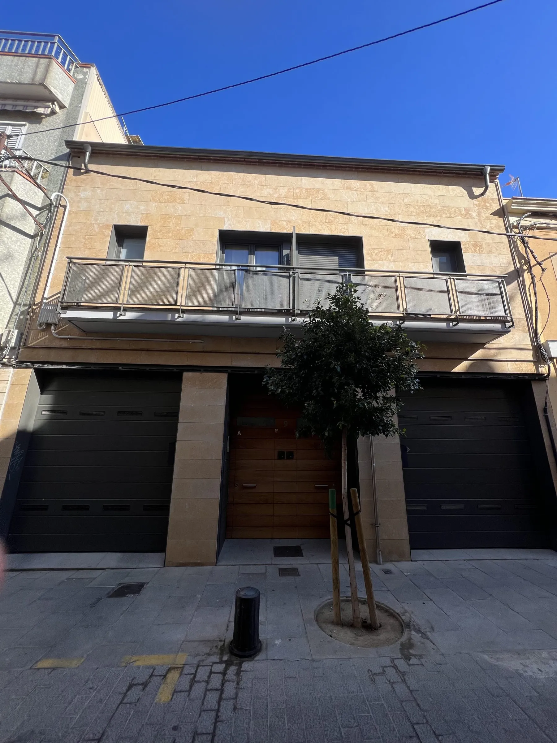 Vista frontal de las dos casas pareadas de Cornellà: piedra clara, barandilla metálica y arbolado en la acera